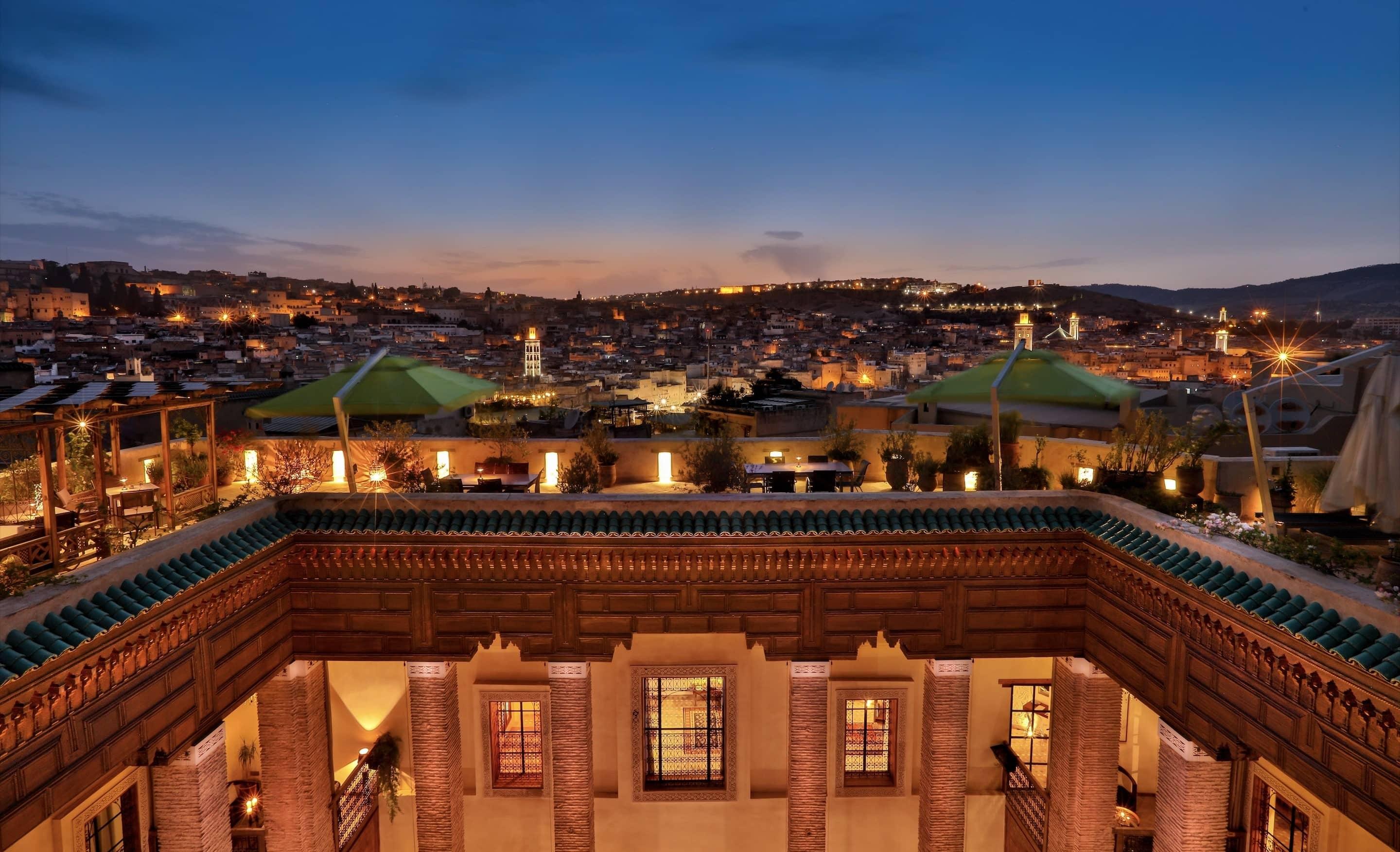 A twilight view from the Karawan Riad, overlooking the ancient city of Fes, with its myriad of lights and historical buildings, all seen from a richly decorated rooftop with Moorish elements.