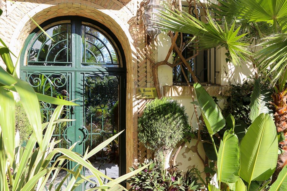 An ornate green door set in a brick archway, framed by tropical plants, marks the entrance to La Maison de Tanger.