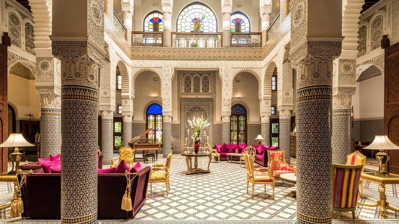 Luxurious Riad Fes' lobby with detailed mosaic floors, grand columns, and colorful stained-glass windows, furnished with magenta sofas and gold-trimmed chairs.