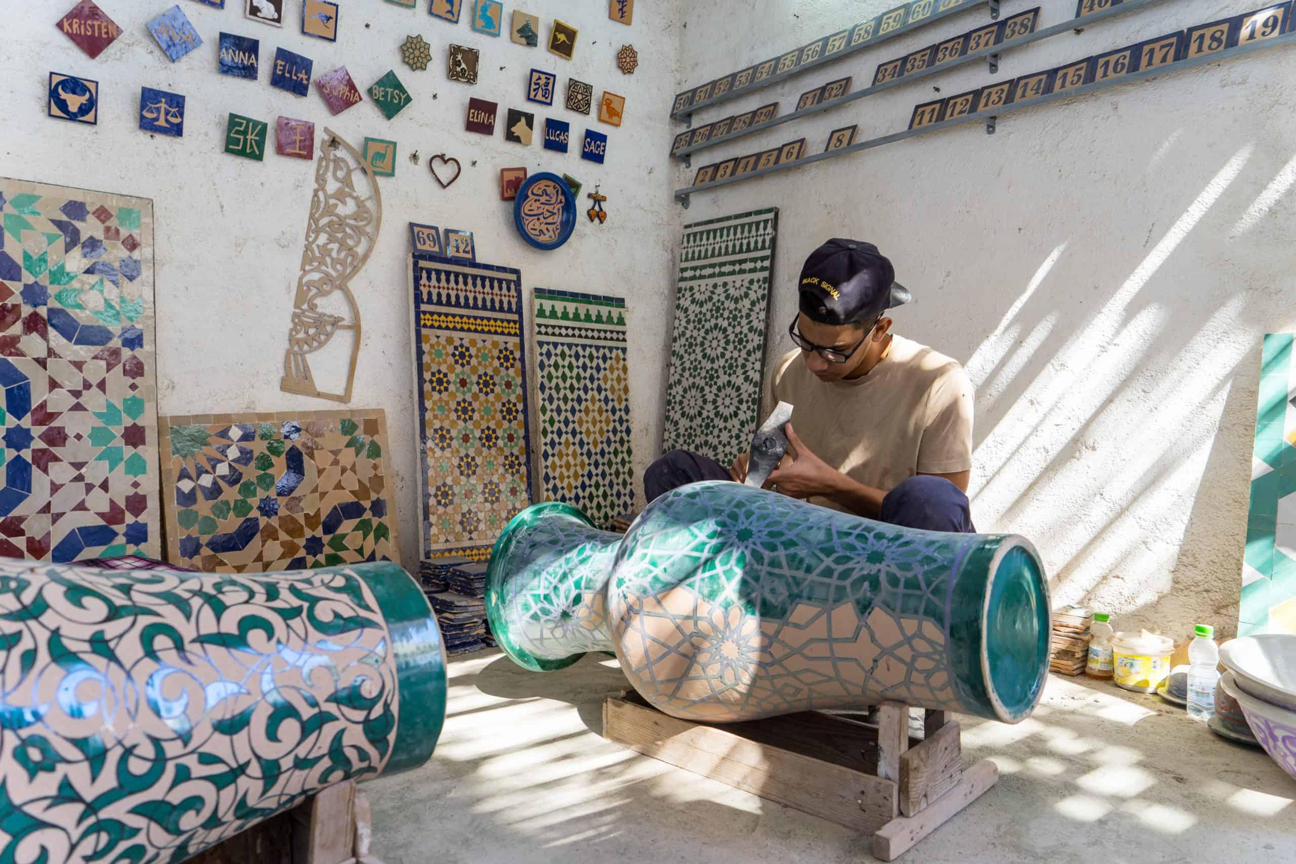 a cooking class on a charming rooftop in Fes, Morocco. Participants, some wearing traditional Moroccan caps, are preparing ingredients at their individual stations surrounded by lush potted plants and vibrant red walls in the background.