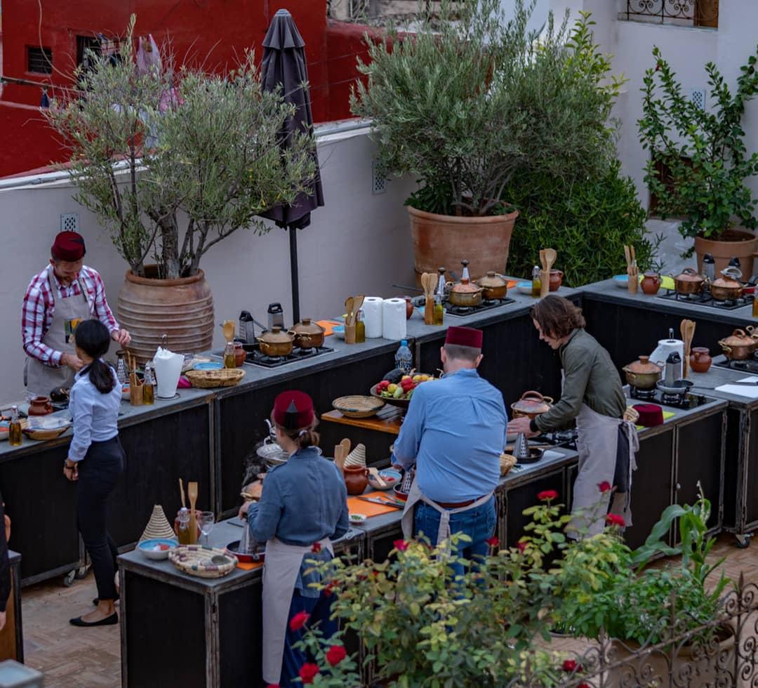 a cooking class on a charming rooftop in Fes, Morocco. Participants, some wearing traditional Moroccan caps, are preparing ingredients at their individual stations surrounded by lush potted plants and vibrant red walls in the background.