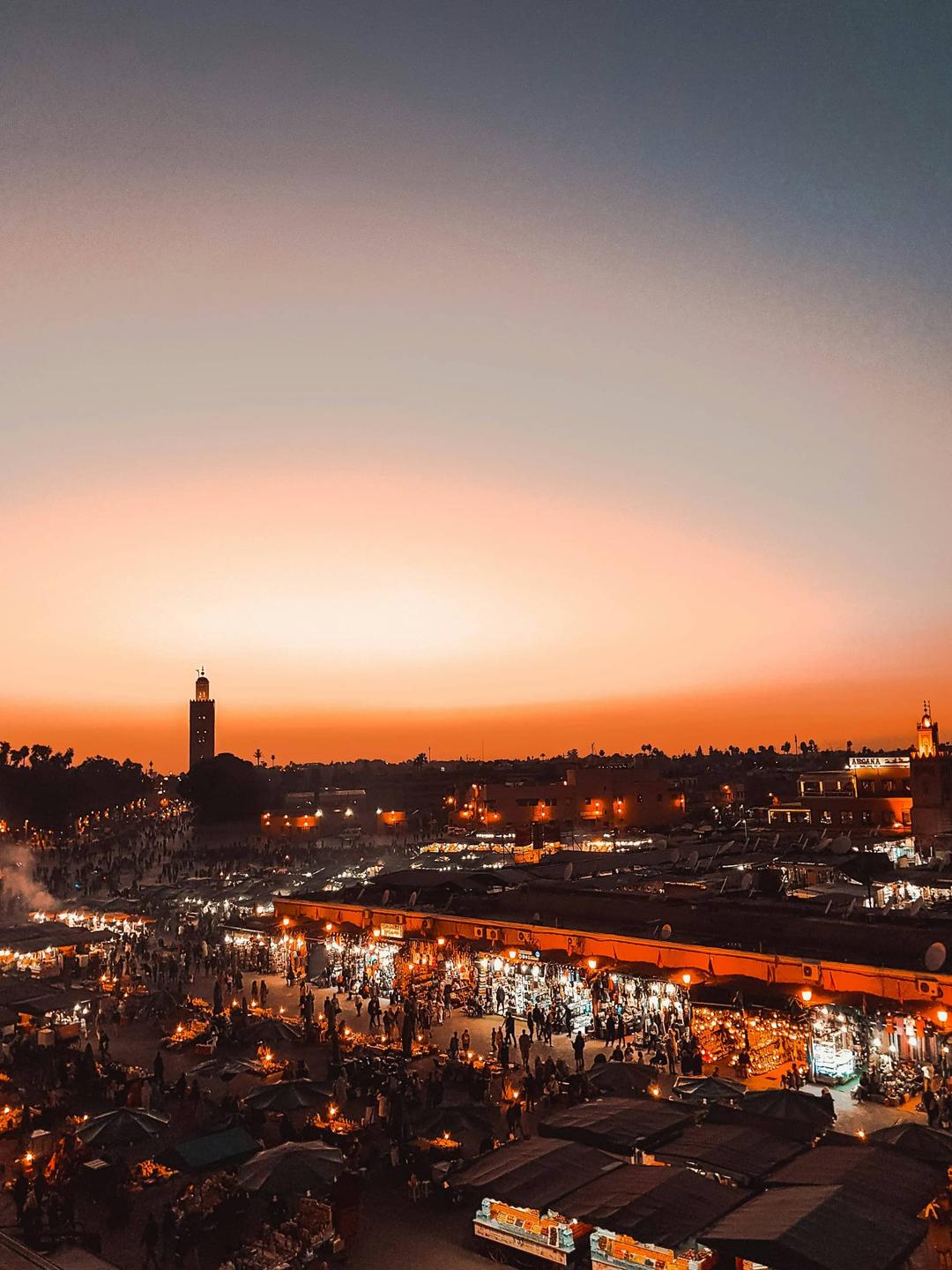 Cover of a luxury travel guide to Morocco featuring an overhead view of the bustling Djemaa el-Fna square in Marrakesh at sunset, with vibrant market stalls, illuminated by warm lights, and the Koutoubia Mosque visible in the background.