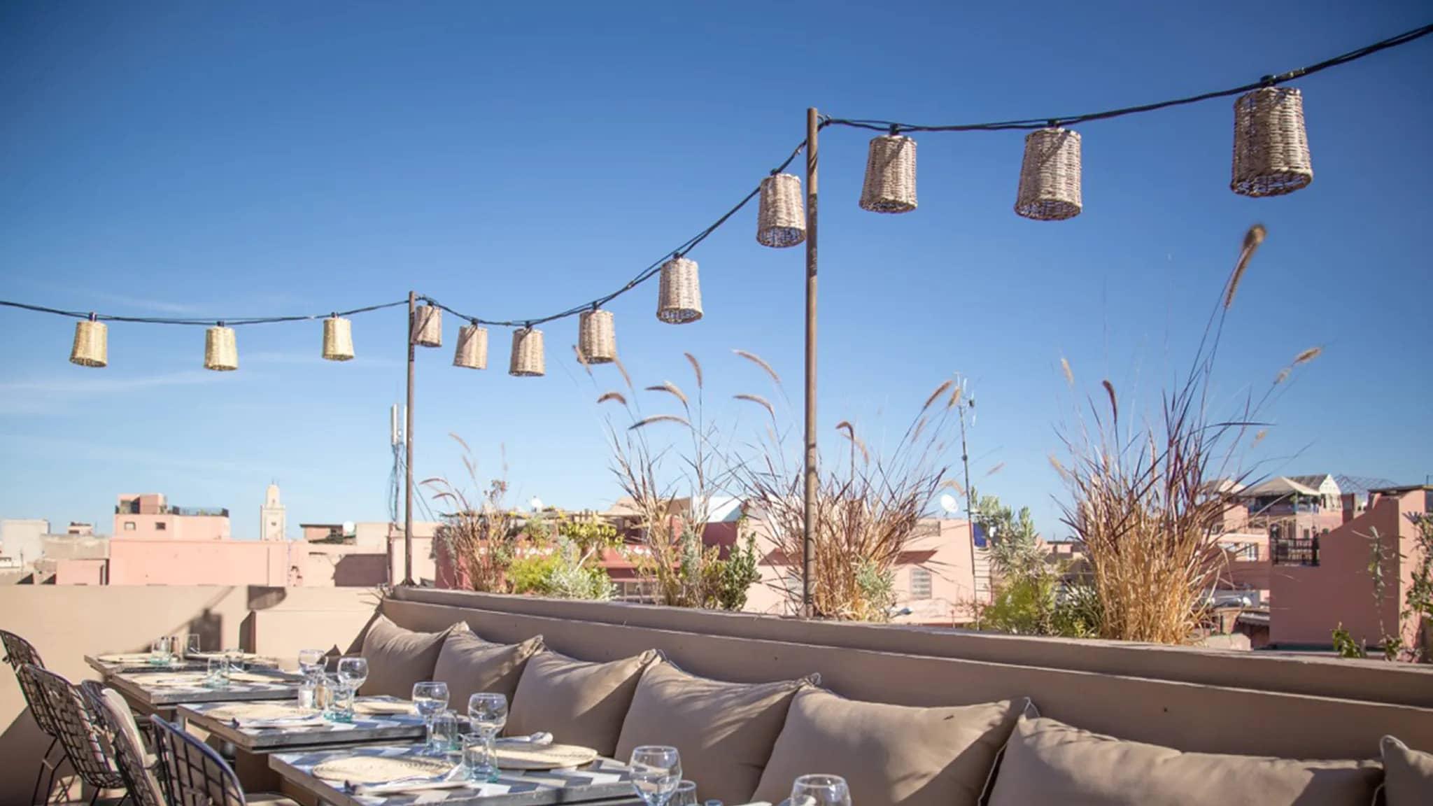 A charming rooftop dining area at Nomad in Marrakech, featuring comfortable seating, ambient hanging lights, and a scenic view of the city's rooftops under a clear blue sky.