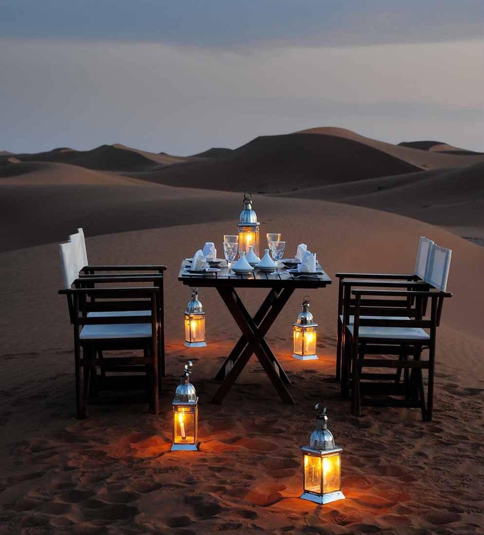 A romantic dining setup at Azalai Desert Camp, featuring a table illuminated by lanterns amidst the serene sand dunes, offering a unique and enchanting experience.