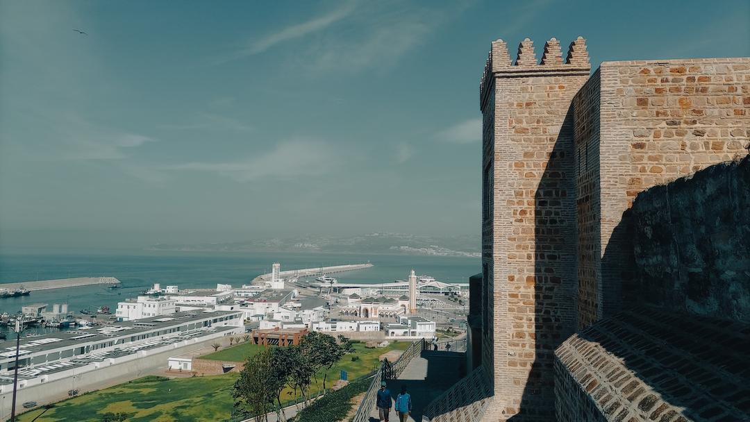 The ancient Kasbah walls of Tangier overlook a modern port with clear views to the coast, under a hazy sky.