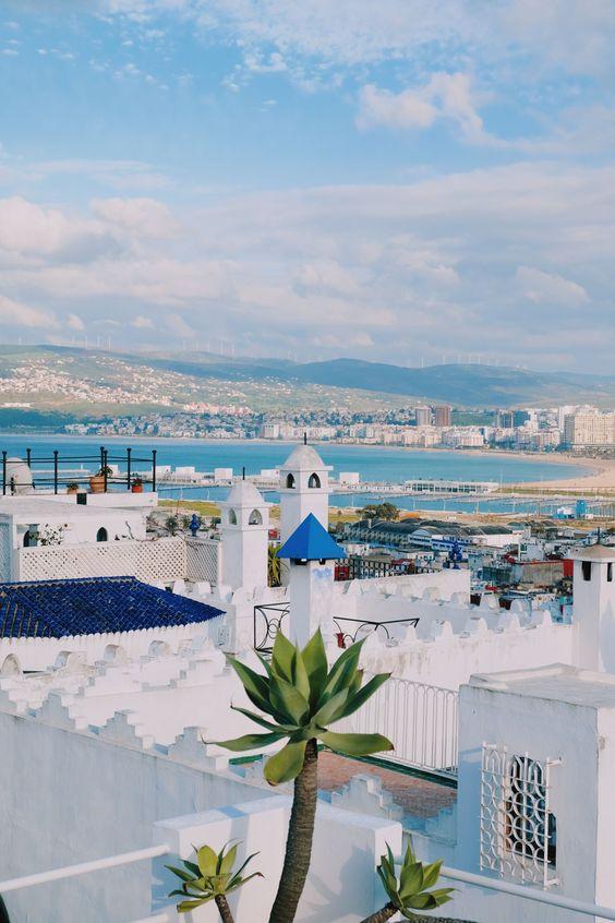 A serene view of Tangier from a rooftop, featuring traditional white buildings with blue accents and a backdrop of the city's coastline and hills.